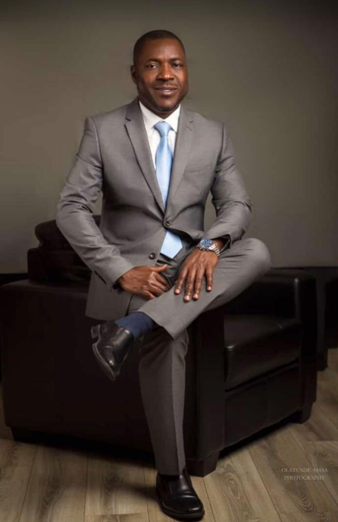 Professional headshot of Alaba Razaq Kokumo, a Black male solicitor wearing a grey business suit with light blue tie, seated confidently on the armrest of a black leather chair in a studio setting with neutral beige and brown tones. He displays a warm, approachable smile while maintaining a professional demeanor.